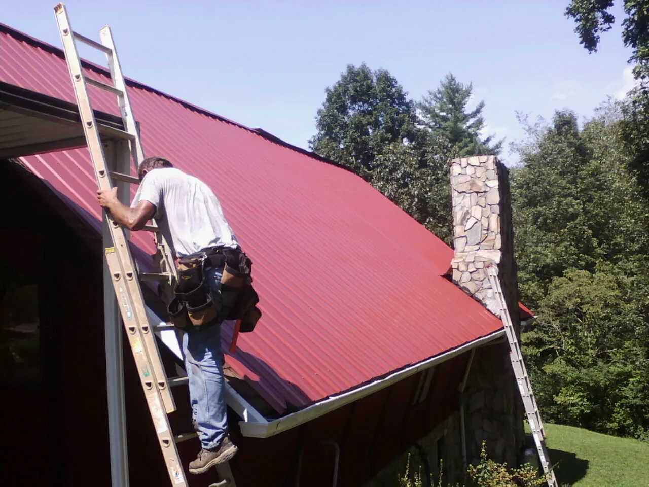 Roofer on a ladder beside a red metal roof and stone chimney — Soffit & Fascia Repair by Roofing Brothers in Bruceton Mills